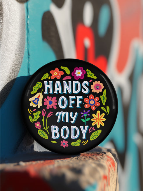 Glossy black pinback button with floral art and bold white text that reads “Hands Off My Body.” Feminist political button featuring bright pastel flowers in pink, red, orange, and yellow. Metal pin with durable safety pin back, shown in various settings: held by hand, displayed on pedestal, and against colorful street art. Available in 2" and 3" sizes, perfect for backpacks, jackets, tote bags, or as a gift for feminist allies and book lovers.